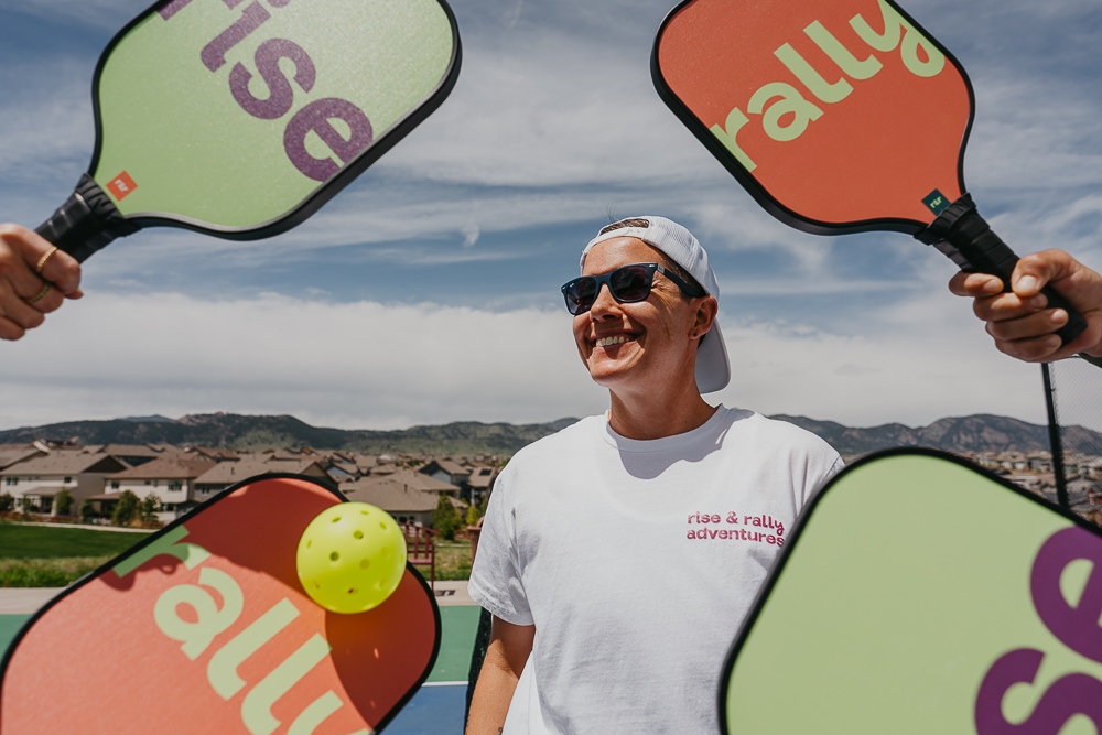 Brand photo of coach smiling with paddles on pickleball court.