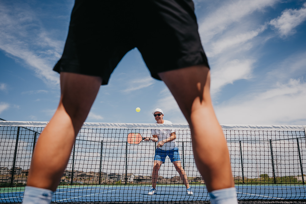 Brand photo coach playing on pickleball court through players legs.